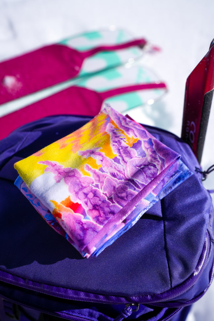 A folded The Snowy Path headband on top of a purple ski backpack with a snowy background